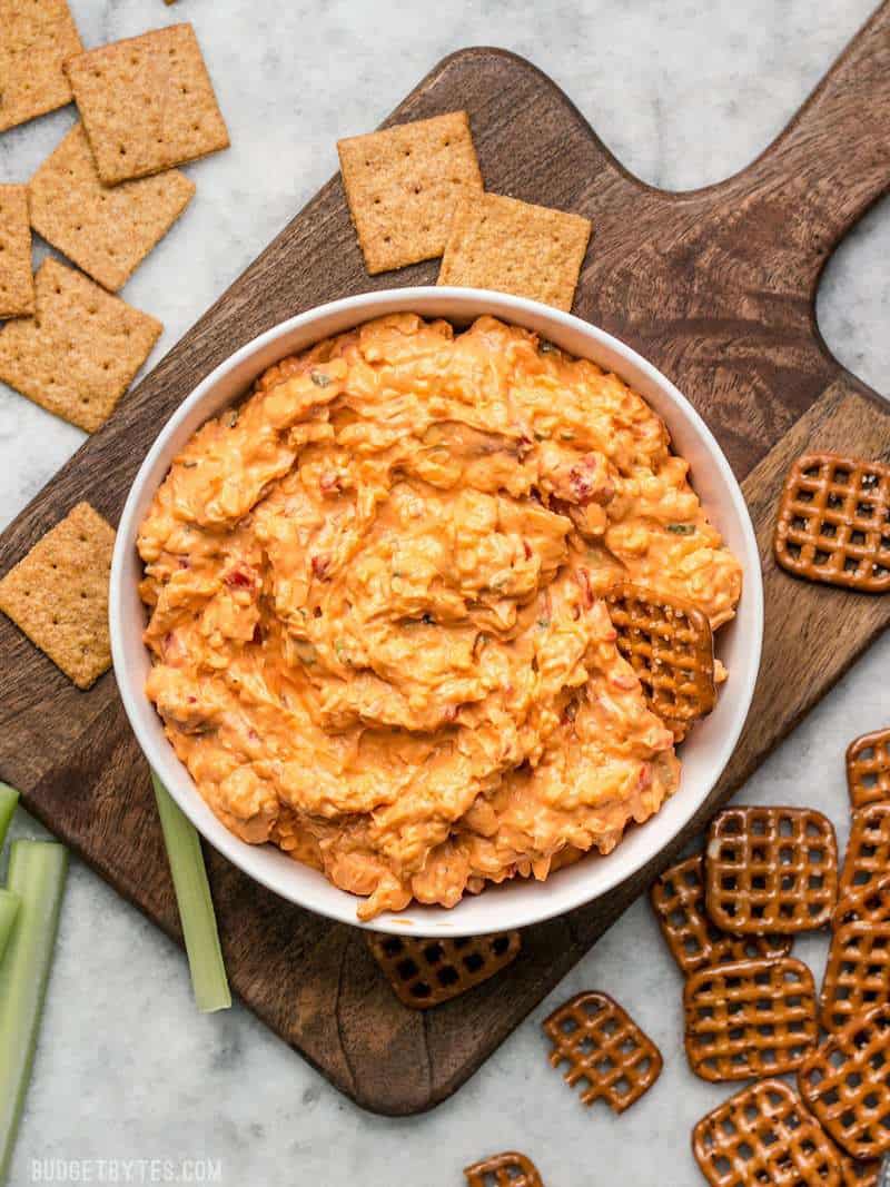 This Sriracha Pimento Cheese Spread is a rich, smoky, spicy spread with just a hint of sweetness from roasted red peppers! BudgetBytes.com A bowl of Sriracha Pimento Cheese Spread on a wooden cutting board surrounded by crackers and pretzels