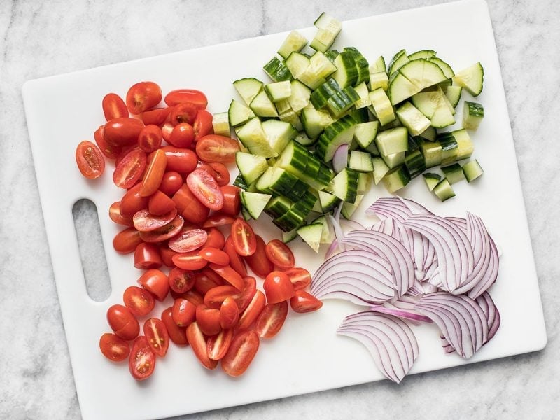 Chopped cucumber, tomato, and onion on a white cutting board.