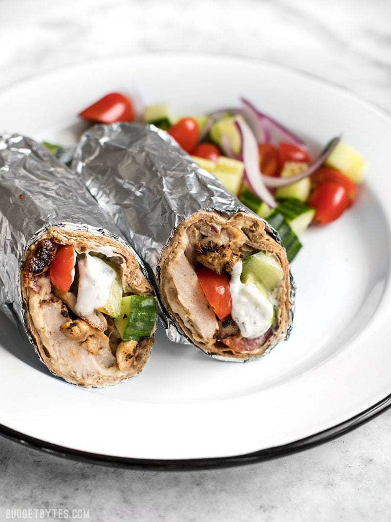 Greek Chicken Wraps wrapped in foil on a plate with the chopped veggies.