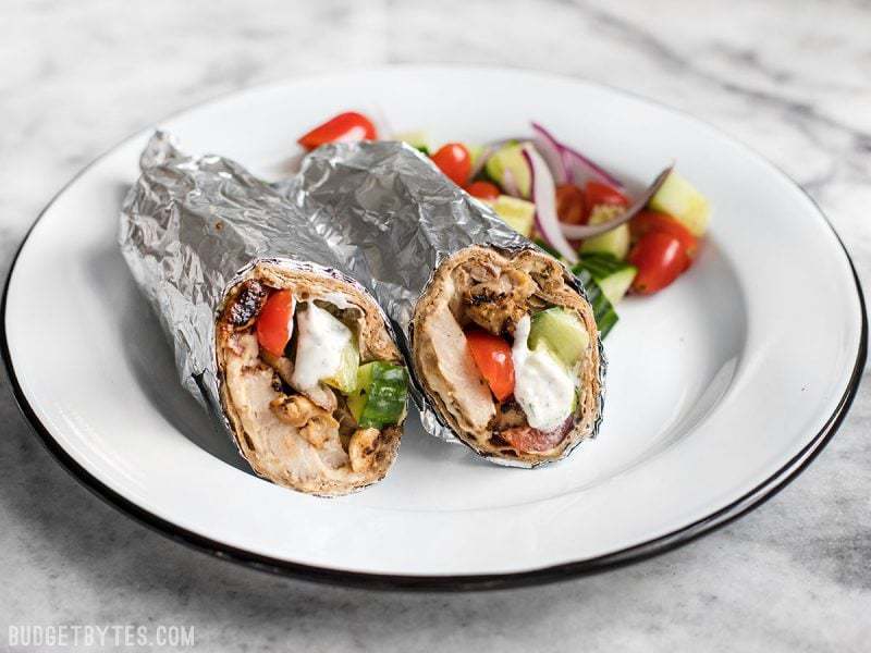 Prep the ingredients for these Greek Chicken Wraps to keep on hand to build fresh wraps or salads all week. BudgetBytes.com Finished Greek Chicken Wraps on a plate with chopped tomatoes, cucumber, and onion.