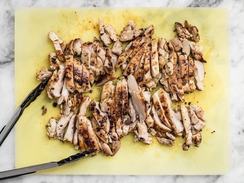 Chicken thighs sliced on a white cutting board with tongs.