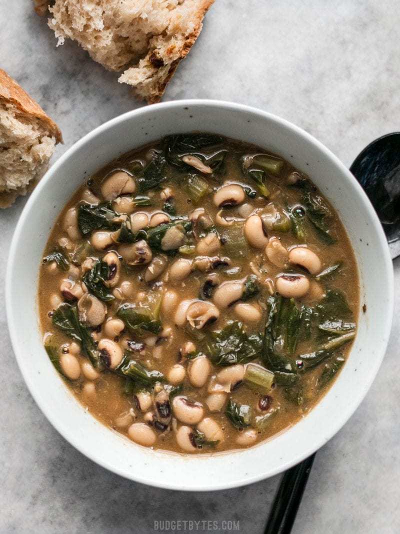 Slow Simmered Black Eyed Peas and Greens is a great cold weather comfort food that is as healthy as it is delicious! Vegan comfort food at its best! Budgetbytes.com A bowl full of Slow Simmered Black Eyed Peas with greens, served with crusty bread.