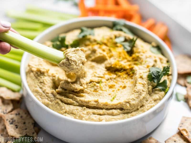 This Coconut Curry Hummus throws a new twist on the traditional dip with creamy coconut milk, vibrant lime, and warm curry spices. BudgetBytes.com A celery stick being dipped into a bowl of Coconut Curry Hummus.
