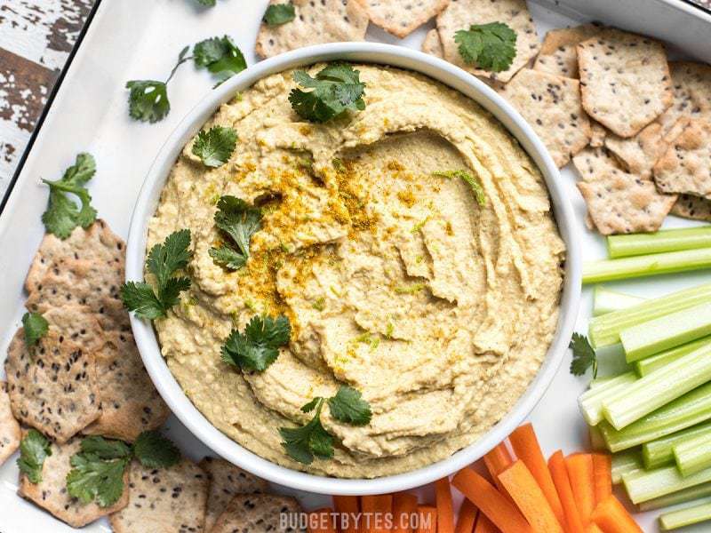 This Coconut Curry Hummus throws a new twist on the traditional dip with creamy coconut milk, vibrant lime, and warm curry spices. BudgetBytes.com Cilantro sprinkled over a bowl of Coconut Curry Hummus surrounded by crackers and vegetable sticks