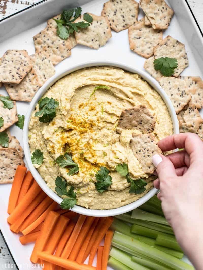 This Coconut Curry Hummus throws a new twist on the traditional dip with creamy coconut milk, vibrant lime, and warm curry spices. BudgetBytes.com A platter of vegetables, crackers, and coconut curry hummus, and a hand dipping a cracker into the hummus.