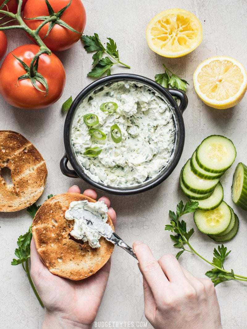 This tangy, herb-infused Scallion Herb Cream Cheese Spread is perfect for sandwiches, wraps, or even snacking on fresh vegetables. BudgetBytes.com Scallion Herb Cream Cheese Spread being smeared onto a toasted bagel
