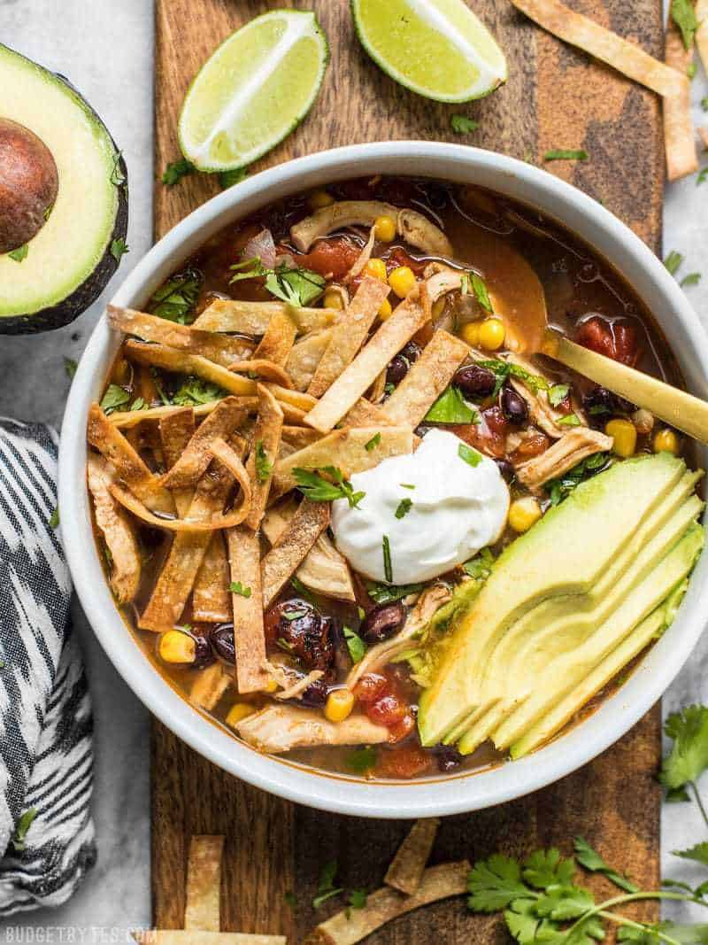 A bowl of Slow Cooker Chicken Tortilla Soup next to limes, avocado, and cilantro