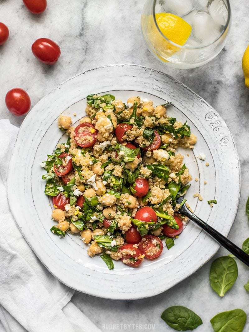 This Spinach Chickpea and Quinoa Salad is an awesome base to build meals throughout the week and it holds up extremely well in the fridge, so you can eat better with less effort. BudgetBytes.com A plate full of Spinach Chickpea and Quinoa Salad with a glass of lemon water, tomatoes, and spinach on the side