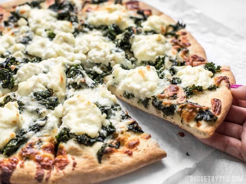 Garlicky greens and creamy ricotta pair perfectly on this light and fresh Garlicky Kale and Ricotta Pizza. It’s the perfect pizza for summer! BudgetBytes.com A hand picking up a piece of Garlicky Kale and Ricotta Pizza