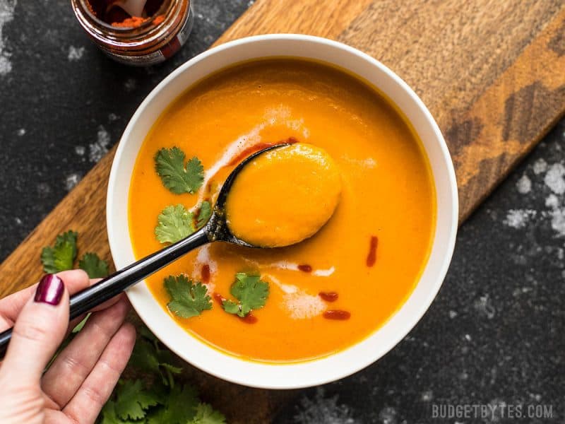 Just a few ingredients are needed make this light, creamy, sweet, and spicy Thai Coconut Curry Carrot Soup. Light, fresh, and delicious! Budgetbytes.com Overhead view of a bowl of Thai Coconut Curry Carrot Soup with a hand lifting a spoonful