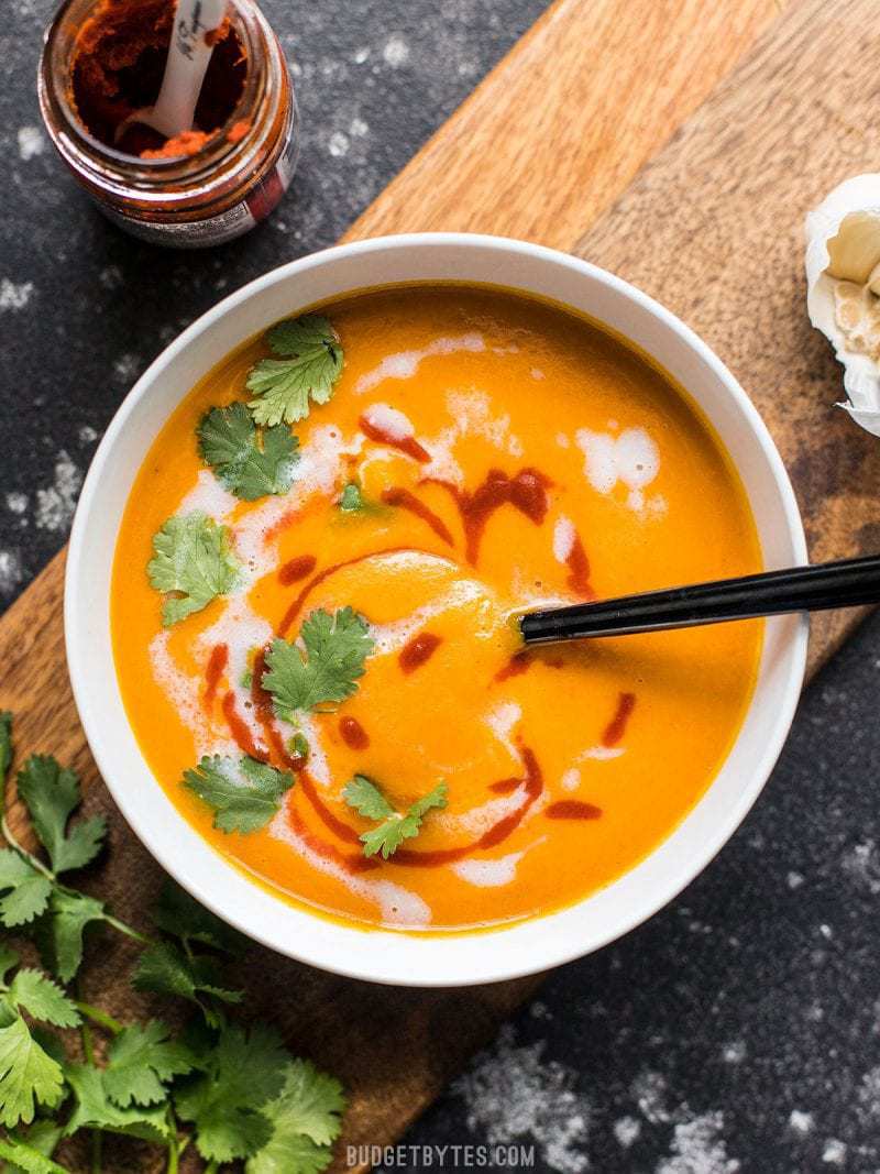 Just a few ingredients are needed make this light, creamy, sweet, and spicy Thai Coconut Curry Carrot Soup. Light, fresh, and delicious! Budgetbytes.com A bowl of Thai Coconut Curry Carrot Soup on a wooden cutting board with coconut milk and sriracha swirled into the top