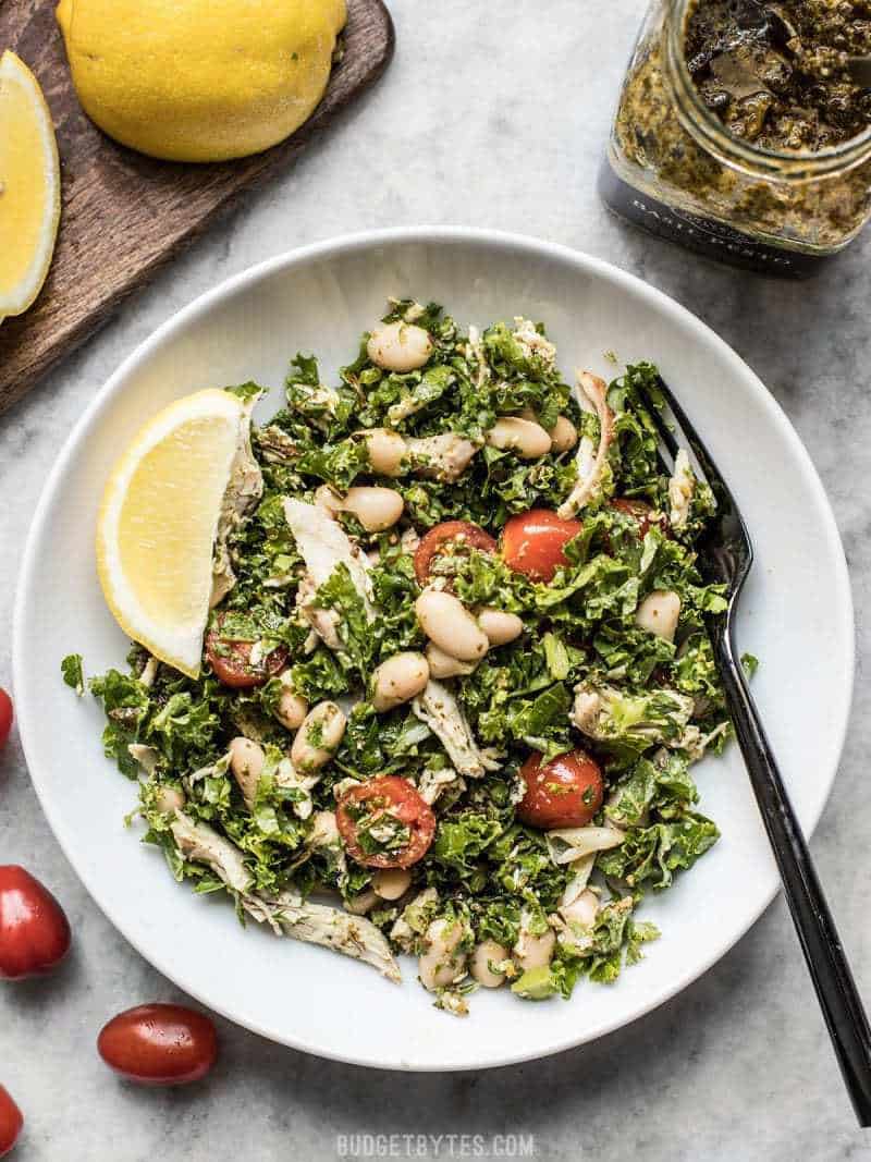 This Make Ahead Kale White Bean and Pesto Salad keeps your fridge stocked with good for you food that you can customize each day for a new meal. Budgetbytes.com A large bowl of Kale White Bean and Pesto Salad with lemon wedges and a black fork.