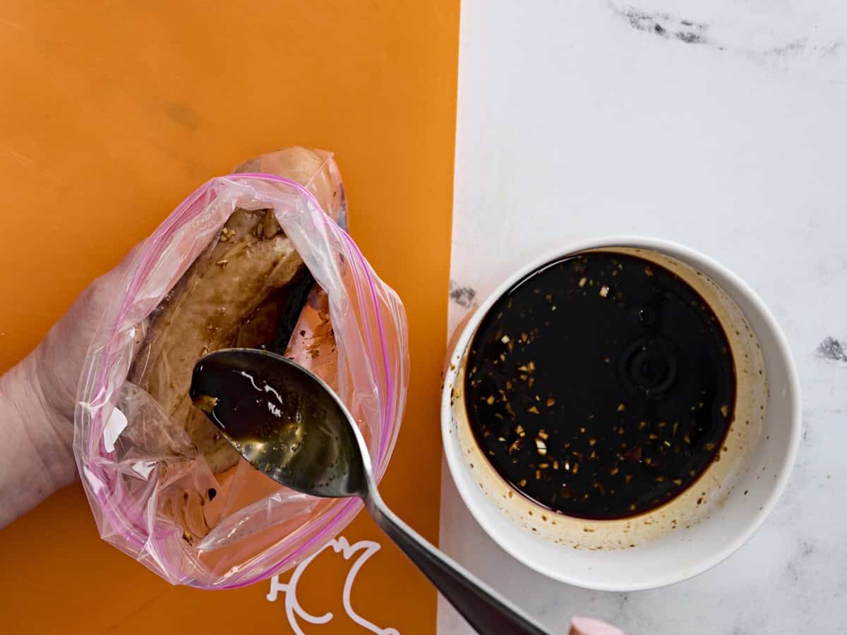 A bowl of homemade teriyaki sauce next to a bag of raw chicken, with the marinade being spooned into the bag.