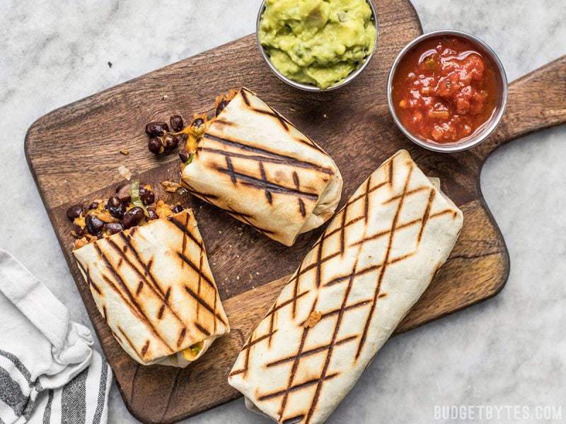 Perfect for stashing in the freezer, these Make Ahead Bean and Cheese Burritos will save you on busy weeknights! Two bean and cheese burritos on a cutting board, one cut in half, with cups of guacamole and salsa on the side.