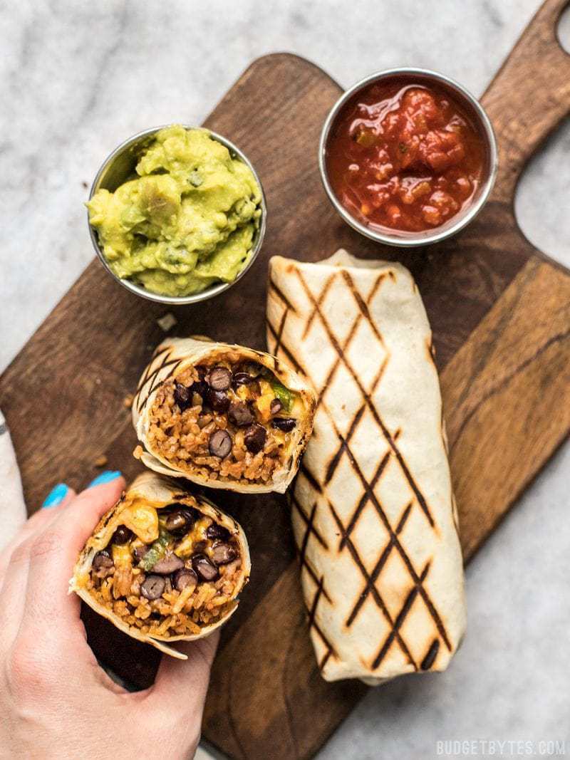 Perfect for stashing in the freezer, these Make Ahead Bean and Cheese Burritos will save you on busy weeknights! A hand holding a bean and cheese burrito ready to eat, with sides of guacamole and salsa.