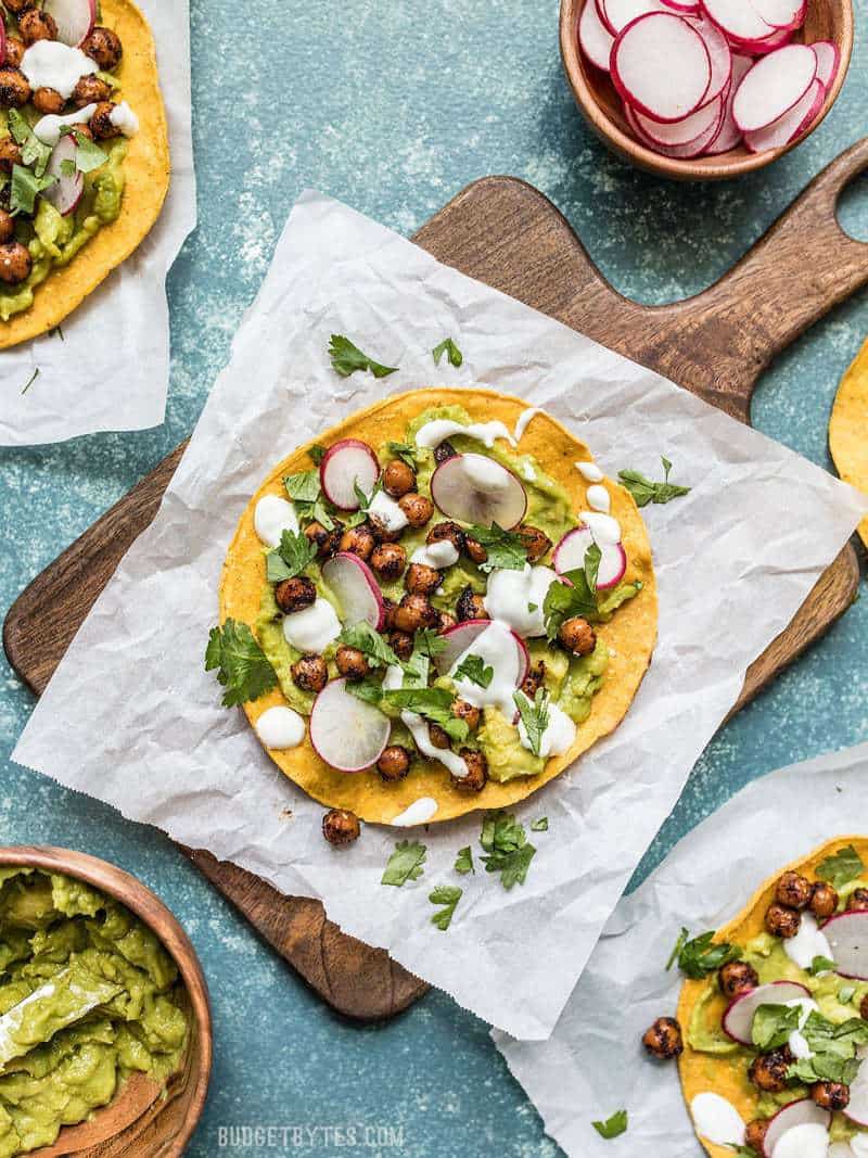 These cool and refreshing Spiced Chickpea Tostadas are the perfect almost no cook dinner for summer. Customize the toppings based on what you have on hand to make the most of leftovers! Budgetbytes.com Spiced Chickpea Tostadas on pieces of parchment and a wooden cutting board