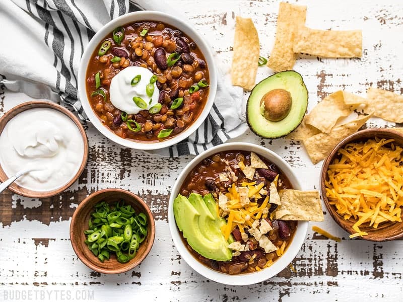 This Slow Cooker Vegetarian Lentil Chili makes a huge batch, is packed with flavor and nutrients, and can be made for only about 5 dollars! Budgetbytes.com Dressed bowls of Slow Cooker Vegetarian Lentil Chili with various toppings on the side