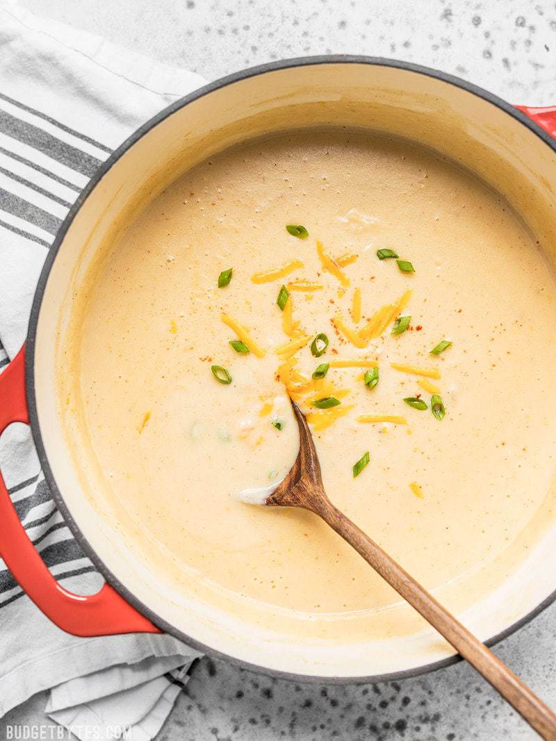 Cheesy Cauliflower and Potato Soup is rich and creamy without being overly heavy. Eat your vegetables and enjoy them, too! Budgetbytes.com A wooden ladle spooning Cheesy Cauliflower and Potato Soup out of the pot.