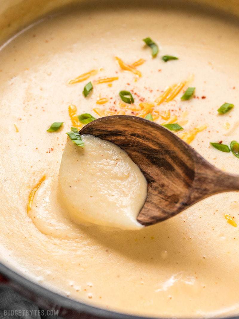 Cheesy Cauliflower and Potato Soup is rich and creamy without being overly heavy. Eat your vegetables and enjoy them, too! Budgetbytes.com Close up of a wooden ladle in a pot of thick and creamy Cheesy Cauliflower and Potato Soup
