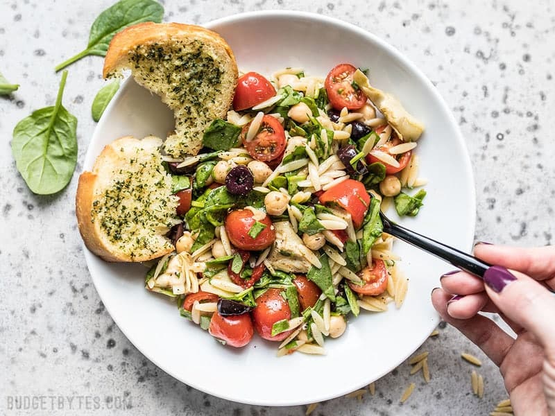 Filling enough to serve as a light meal, but light enough to act as an easy side dish, this Italian Orzo Salad is a versatile addition to your weekly meal prep. Budgetbytes.com A hand taking a forkful of Italian Orzo Salad from a plate. Garlic Bread on the side.
