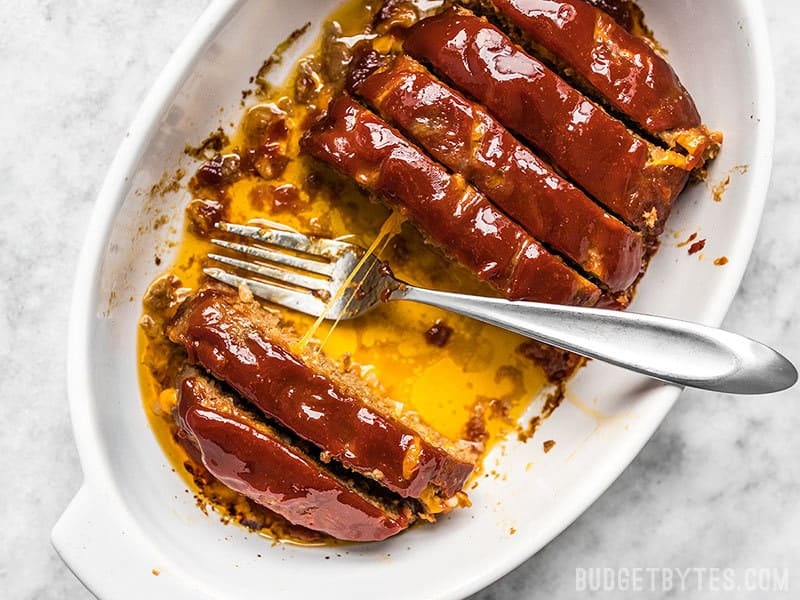 This super cheesy Cheddar Cheeseburger Meatloaf is studded with melty nuggets of cheddar, and topped with a tangy tomato-mustard sauce. Budgetbytes.com Sliced Cheddar Cheeseburger Meatloaf with pulled cheese in a casserole dish with a few slices missing.