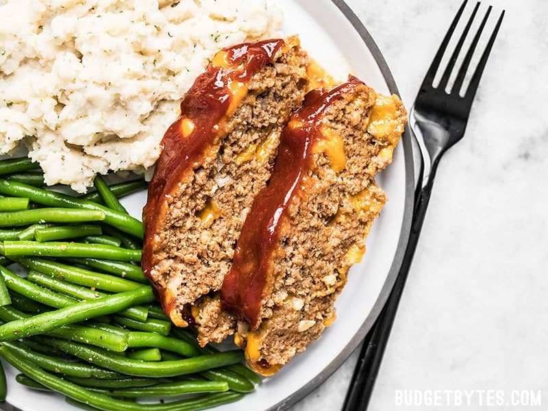 This super cheesy Cheddar Cheeseburger Meatloaf is studded with melty nuggets of cheddar, and topped with a tangy tomato-mustard sauce. Budgetbytes.com Two slices of Cheddar Cheeseburger Meatloaf on a dinner plate with mashed potatoes and green beans.