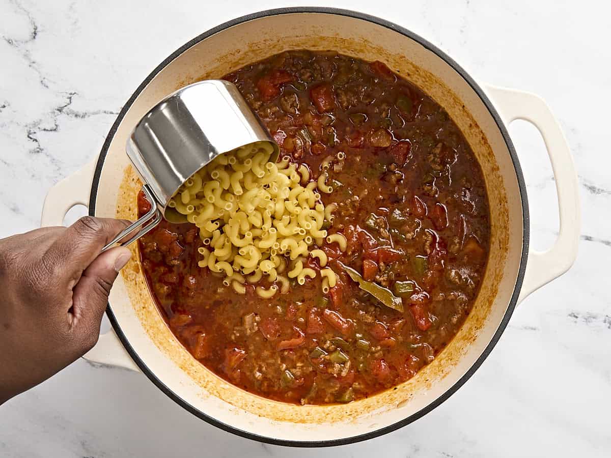 Dried macaroni being added to a soup pot of american goulash.