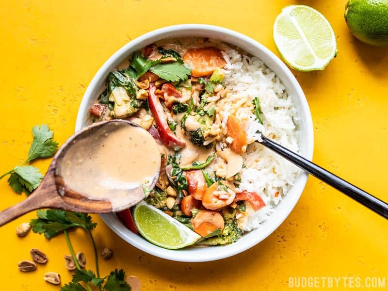 This rich and spicy coconut vegetable stir fry is adaptable to whatever vegetables are lingering in your fridge, making it a great sweep the kitchen recipe! Budgetbytes.com Spooning Spicy Coconut Sauce over a bowl of Spicy Coconut Vegetable Stir Fry and rice