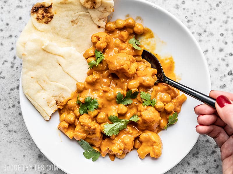 This super easy, ultra creamy, and heavily spiced Cauliflower and Chickpea Masala will be your new favorite weeknight dinner! So much flavor, so little effort. Budgetbytes.com A bowl of creamy Cauliflower and Chickpea Masala being eaten with a spoon and two pieces of naan on the side.