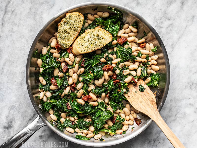 This quick Sun Dried Tomato, Kale, and White Bean Skillet is a fast, flavorful, and fiber-licious meal that is ready for perfect for weekly meal prepping. Budgetbytes.com Finished Sun Dried Tomato, Kale, and White Bean Skillet with wooden spatula