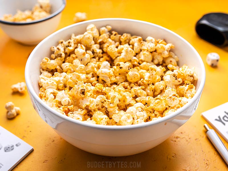 Sriracha Nooch Popcorn is the perfect salty snack with tons of crunch, a spicy buttery coating, and a slightly cheesy flavor thanks to nutritional yeast. Budgetbytes.com Front view of a bowl of Sriracha Nooch Popcorn on a yellow background.