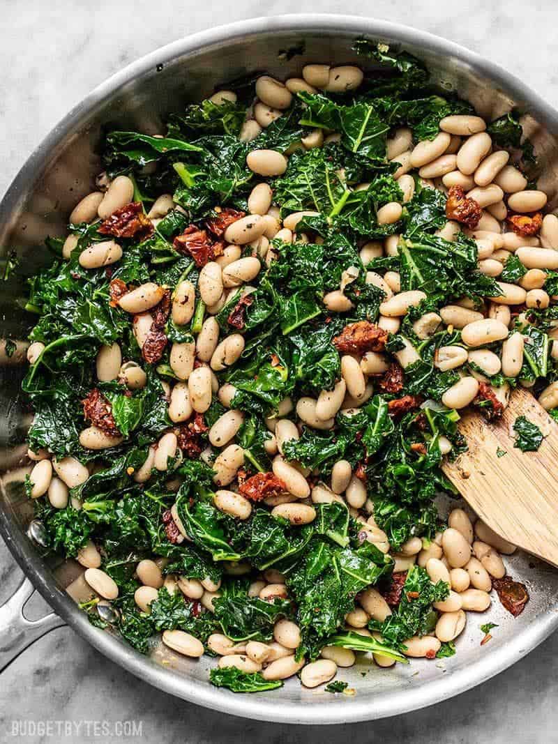 This quick Sun Dried Tomato, Kale, and White Bean Skillet is a fast, flavorful, and fiber-licious meal that is ready for perfect for weekly meal prepping. Budgetbytes.com A skillet full of Sun Dried Tomato, Kale, and White Beans, with a wooden spatula.