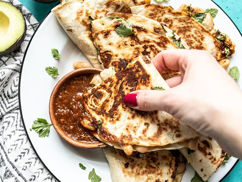Crispy on the outside, creamy on the inside, these insanely delicious Creamy White Bean and Spinach Quesadillas make an awesomely fast weeknight dinner. Budgetbytes.com A hand dipping a piece of creamy white bean and spinach quesadilla into a bowl of red salsa