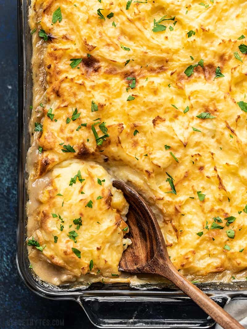 This Cheesy Cottage Pie is chock-full of vegetables and a rich beef gravy, then topped with a layer of super smooth and creamy cheddar mashed potatoes. Budgetbytes.com Close up overhead view of Cheesy Cottage Pie being scooped out of the glass casserole dish with a large wooden spoon.