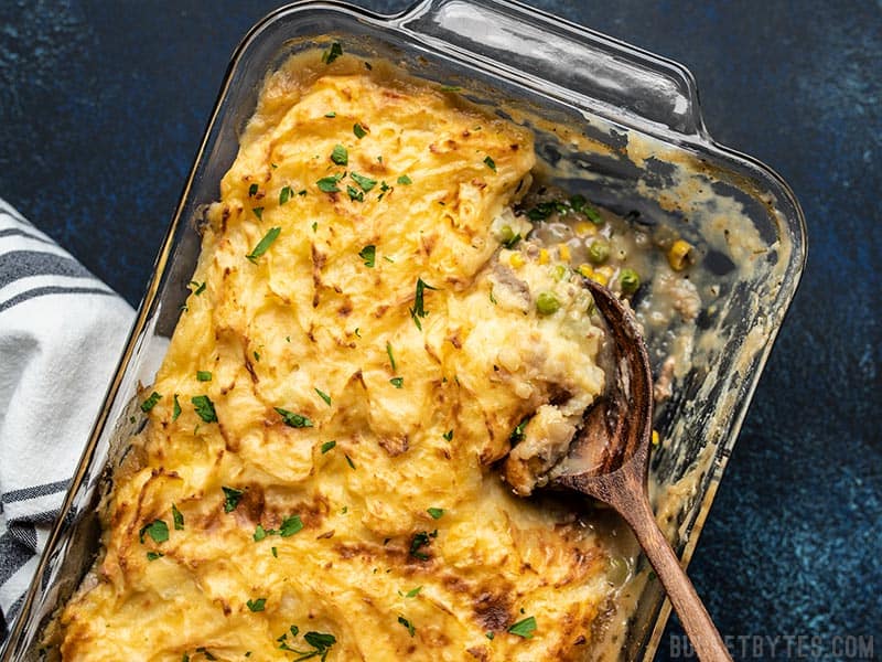 This Cheesy Cottage Pie is chock-full of vegetables and a rich beef gravy, then topped with a layer of super smooth and creamy cheddar mashed potatoes. Budgetbytes.com Cheesy Cottage Pie being scooped out of a glass casserole dish with a big wooden spoon.