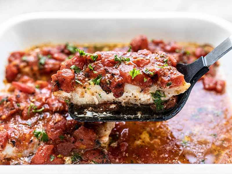 This Easy Oven Baked Fish with Tomatoes is baked in a garlic and herb infused tomato sauce that helps keep it tender and moist as it bakes. Budgetbytes.com A fillet of baked fish with tomatoes being lifted out of the baking dish with a spatula