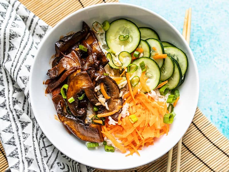 Gochujang Mushroom Bowls feature sweet and spicy marinated mushrooms, quick pickled cucumbers, crunchy carrots, and fragrant jasmine rice.Budgetbytes.com Overhead view of a finished Gochujang Mushroom Bowl on a bamboo mat with chopsticks on the side.