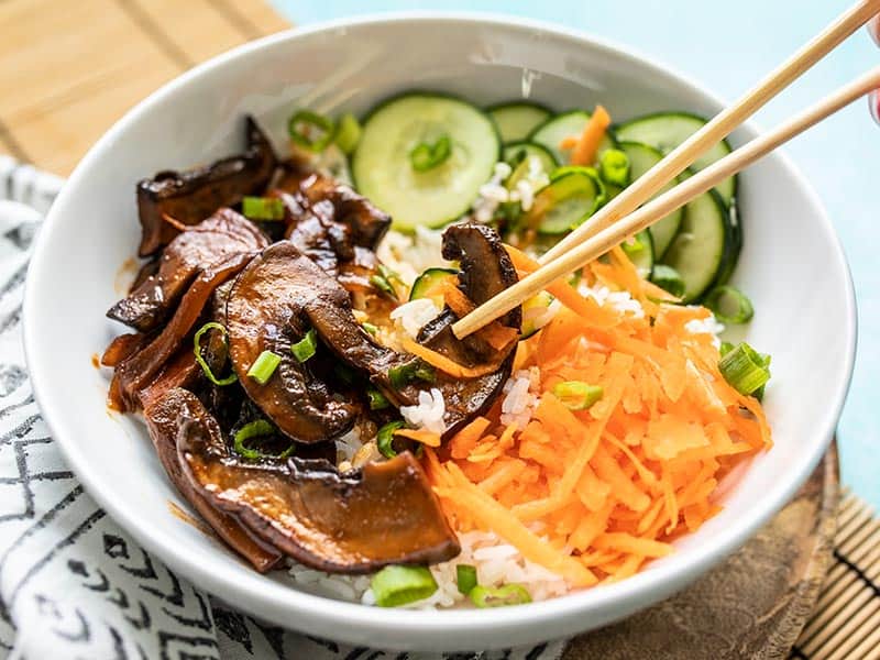 Gochujang Mushroom Bowls feature sweet and spicy marinated mushrooms, quick pickled cucumbers, crunchy carrots, and fragrant jasmine rice.Budgetbytes.com Front view of chopsticks picking up a slice of mushroom out of a Gochujang Mushroom Bowl.