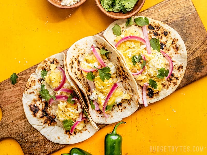 Hummus Breakfast Tacos are a quick and easy, protein and fiber packed breakfast that will keep you fueled all morning. Customize the toppings as desired! Budgetbytes.com Three Hummus Breakfast Tacos on a wooden cutting board with cilantro, cheese, and jalapeños on the sides