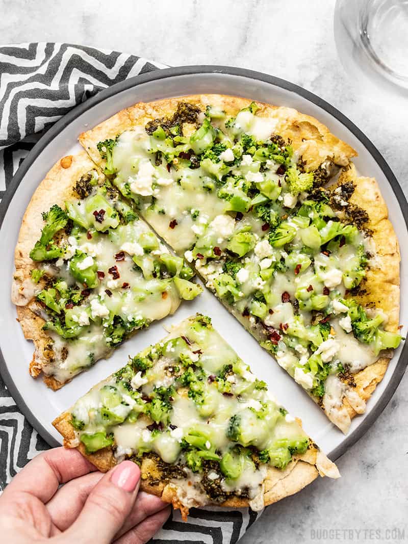 This "Quick Fix" Broccoli Pesto Pizza is the fastest and easiest route to getting your pizza fix, with an extra thin and crispy tortilla crust. Budgetbytes.com A Quick Fix Broccoli Pesto Pizza on a plate with a striped napkin and a hand pulling one slice away.