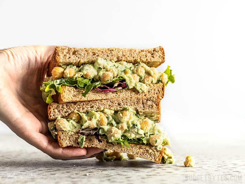 Light, bright, and full of spring flavors, this Scallion Herb Chickpea Salad will keep you full all afternoon. Serve as a sandwich, wrap, or salad! Budgetbytes.com A hand grabbing a stack of two halves of a Scallion Herb Chickpea Salad sandwich on wheat bread.