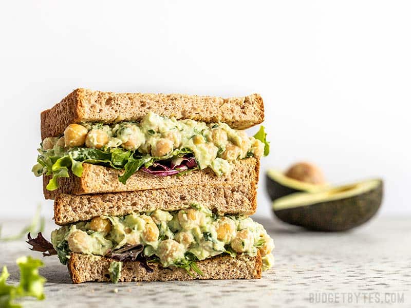 Light, bright, and full of spring flavors, this Scallion Herb Chickpea Salad will keep you full all afternoon. Serve as a sandwich, wrap, or salad! Two halves of a Scallion Herb Chickpea Salad sandwich on wheat bread, stacked. An avocado in the background.
