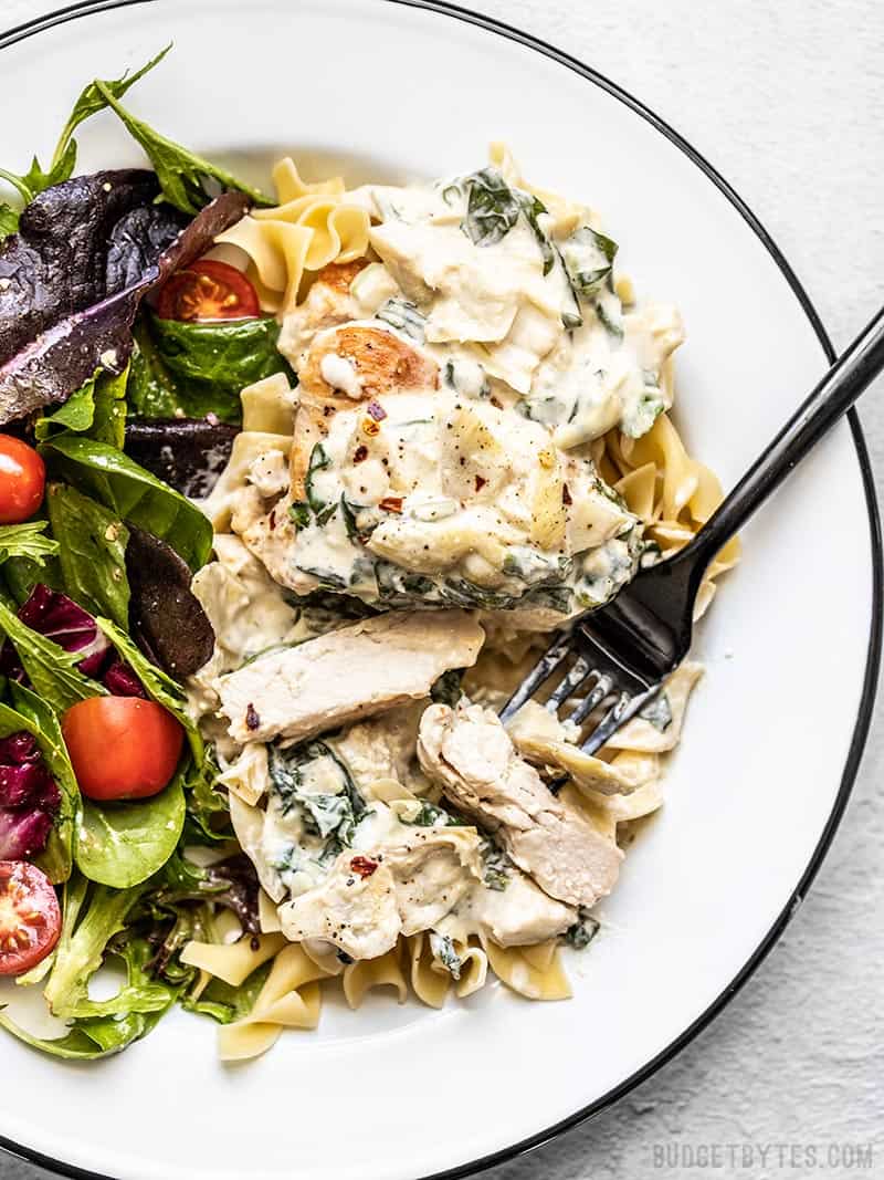 Creamy Spinach Artichoke Chicken is a creamy and indulgent chicken dish that is fast and easy, but still impressive enough for date night. Budgetbytes.com Creamy Spinach Artichoke Chicken on a plate with a bed of pasta and a salad on the side