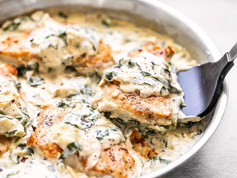 Creamy Spinach Artichoke Chicken is a creamy and indulgent chicken dish that is fast and easy, but still impressive enough for date night. Budgetbytes.com A piece of Creamy Spinach Artichoke Chicken being lifted out of the skillet with a spatula, side view