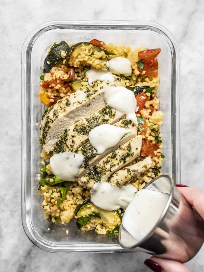 Roasted Vegetable Couscous Meal Prep is a super filling, veggie-packed meal that can be made with chicken or vegetarian with chickpeas. BudgetBytes.com A glass meal prep container with roasted vegetable couscous and sliced chicken, with ranch being drizzled over top.