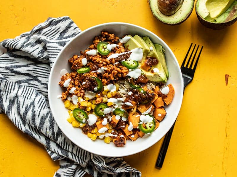 Spiced and crumbled tempeh makes a fast and easy alternative to beef in these Tempeh Burrito Bowls. Customize the toppings to be vegetarian or vegan! Budgetbytes.com Overhead view of a finished tempeh burrito bowl on a yellow background with a black and white napkin and an avocado on the side.