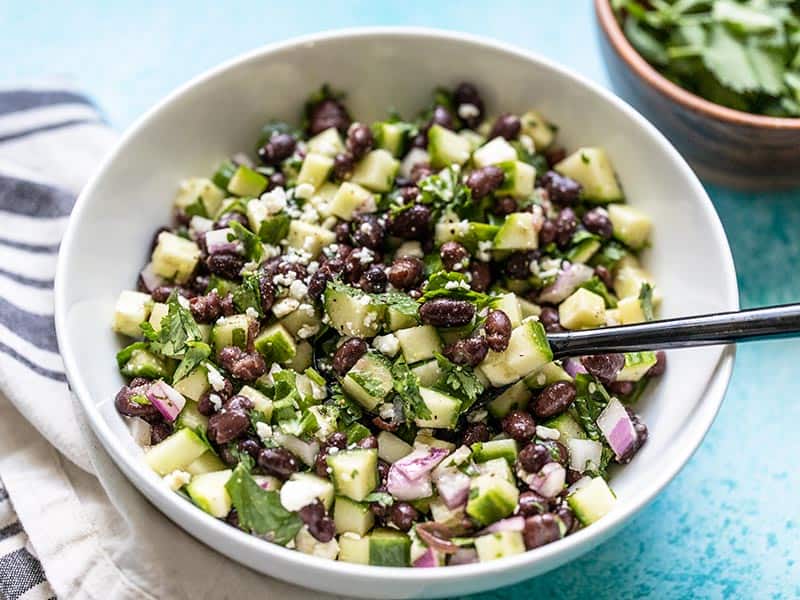 This light and fresh cucumber and black bean salad is the perfect summer side dish. It is refrigerator stable so make it once and snack on it all week! Budgetbytes.com Front view of Cucumber and Black Bean Salad in a white bowl with a black spoon.