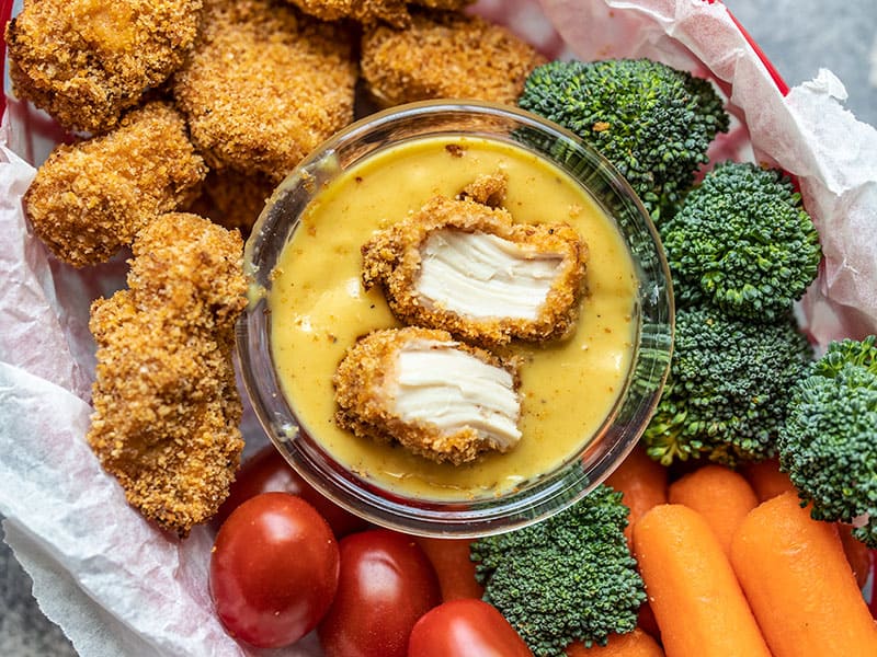 Homemade baked chicken nuggets are crispy on the outside, tender and juicy on the inside, easy to make, and make a delicious meal or snack. Budgetbytes.com One chicken nugget ripped in half, sitting in a bowl of honey mustard, surrounded by vegetables and nuggets.