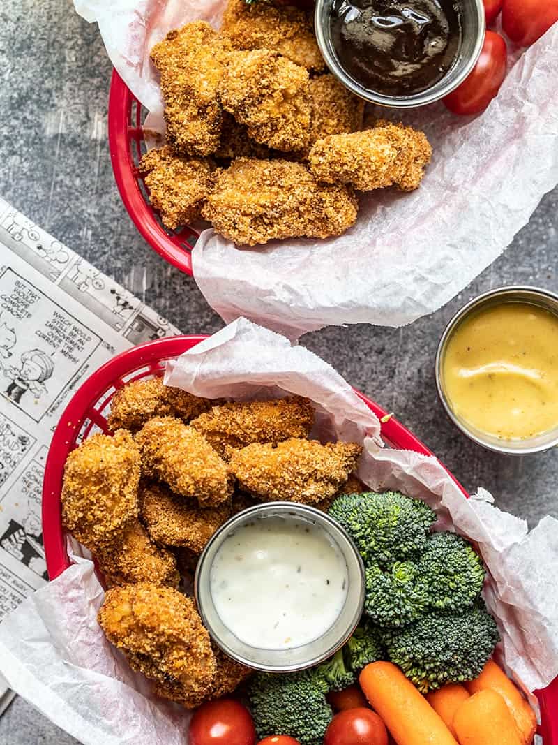 Homemade baked chicken nuggets are crispy on the outside, tender and juicy on the inside, easy to make, and make a delicious meal or snack. Budgetbytes.com Two baskets with Homemade Baked Chicken Nuggets, vegetables, and dip sitting next to a newspaper.