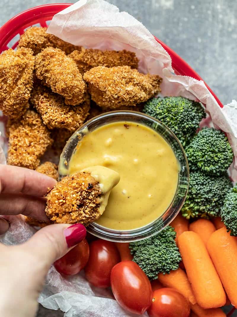 Homemade baked chicken nuggets are crispy on the outside, tender and juicy on the inside, easy to make, and make a delicious meal or snack. Budgetbytes.com One Homemade Baked Chicken Nugget being dipped into honey mustard, surrounded by more nuggets and vegetables.